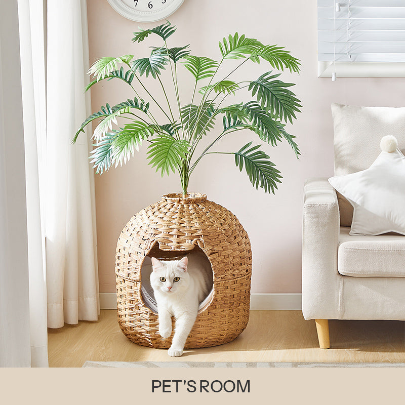 Woven rattan cat house cave topped with an artificial green palm plant, with a white cat looking out inside a living room.