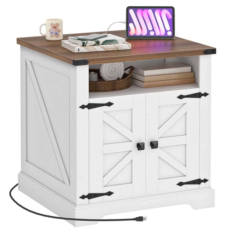 A white farmhouse-style side table with a brown wood top, featuring two barn-door cabinet doors with black hardware, an open shelf with books and a basket, and a tablet on the desktop. A mug sits on the left side, and a power cord is visible in the foreground against a plain white background.