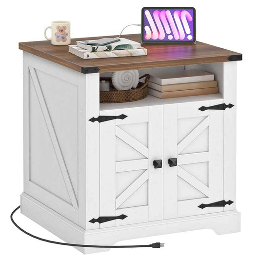 A white farmhouse-style side table with a brown wood top, featuring two barn-door cabinet doors with black hardware, an open shelf with books and a basket, and a tablet on the desktop. A mug sits on the left side, and a power cord is visible in the foreground against a plain white background.