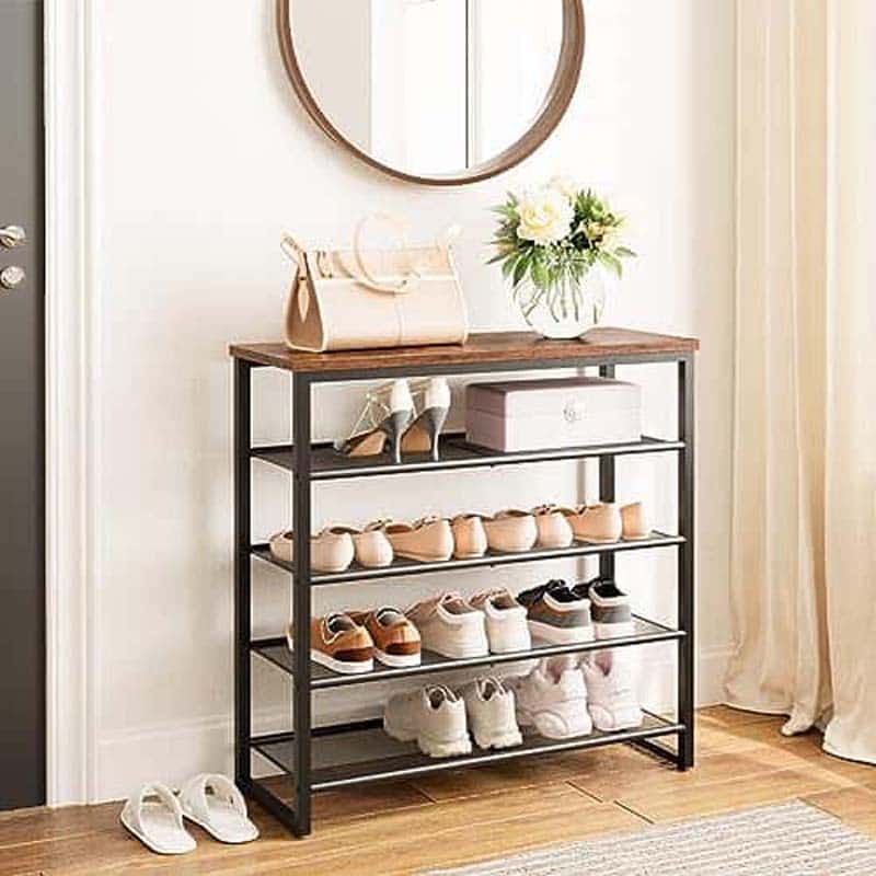 A 4-tier shoe rack with a rustic dark wood top and black metal frame, styled with various shoes, a handbag, and a vase of flowers in a bright entryway.