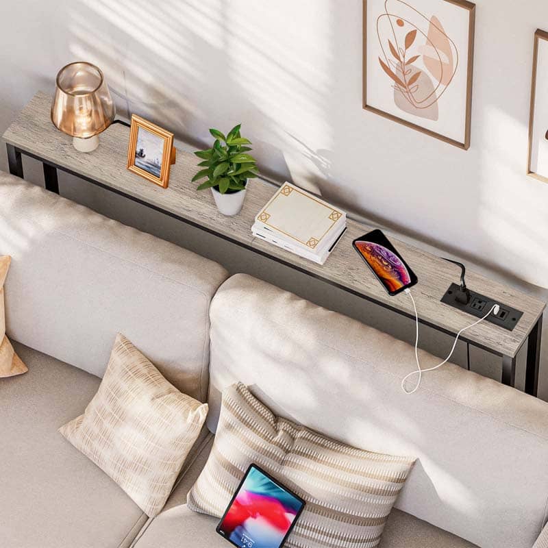 A modern console table with a gray wood-grain top and black metal frame, styled in a living room setting on a beige sofa. The table features built-in side outlets with a black power strip, a copper-colored table lamp, a small potted green plant, a notebook, and a smartphone.