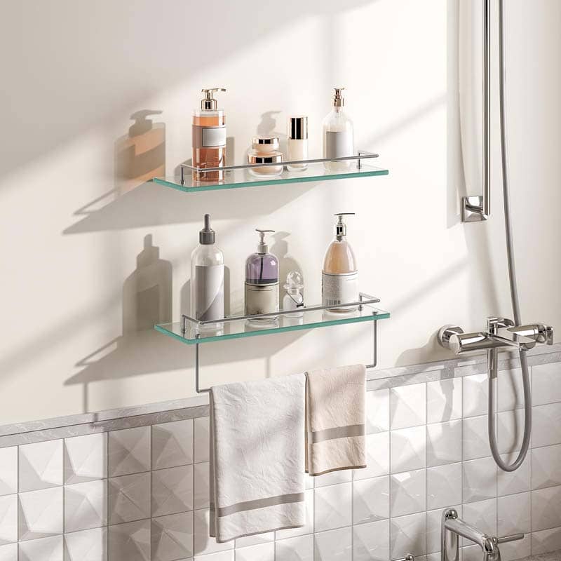 Two clear glass wall-mounted shelves with silver metal brackets holding assorted bathroom toiletries, installed on a white tiled wall above a bathtub with a hanging towel.