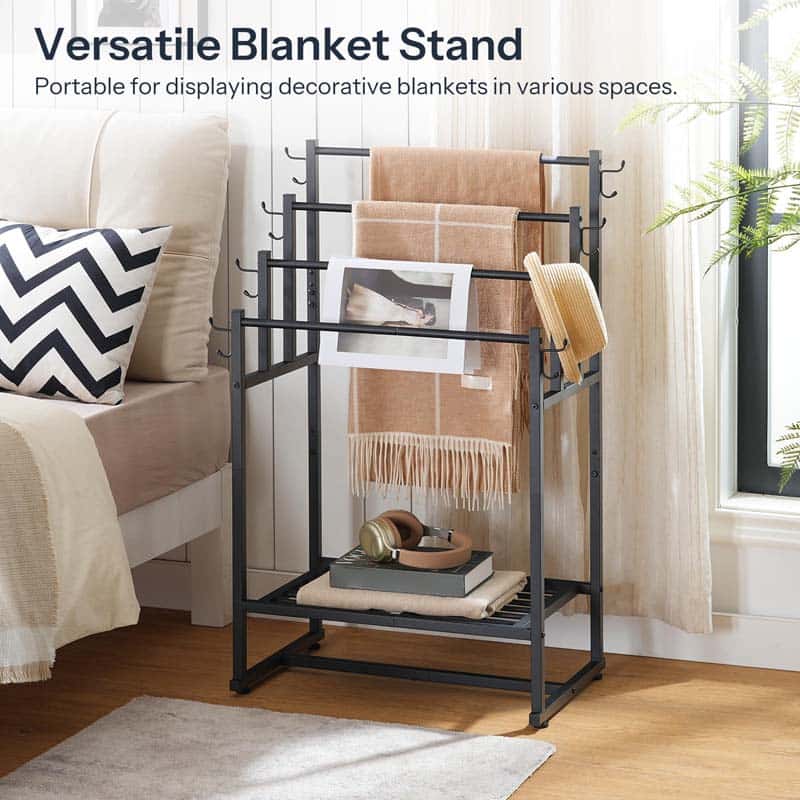A black metal versatile blanket stand in a bedroom setting, holding a beige fringed throw blanket, a book, and a hat. The text at the top reads 'Versatile Blanket Stand Portable for displaying decorative blankets in various spaces.