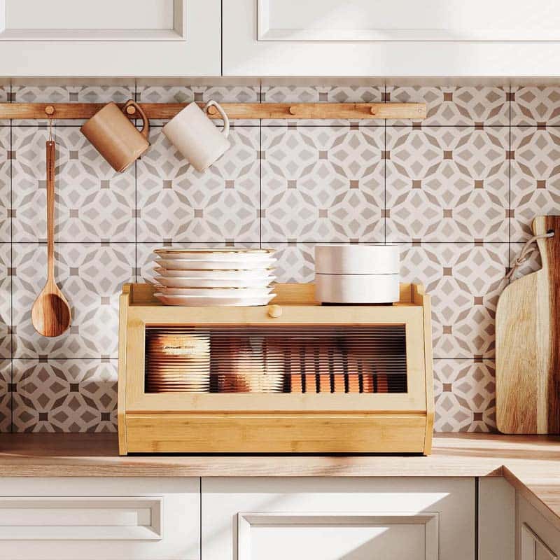 A light natural wood dish rack with a glass-front door, shown on a kitchen counter against a patterned tile backsplash. White dishes are stacked on top, and kitchen utensils hang on a wooden rack behind.