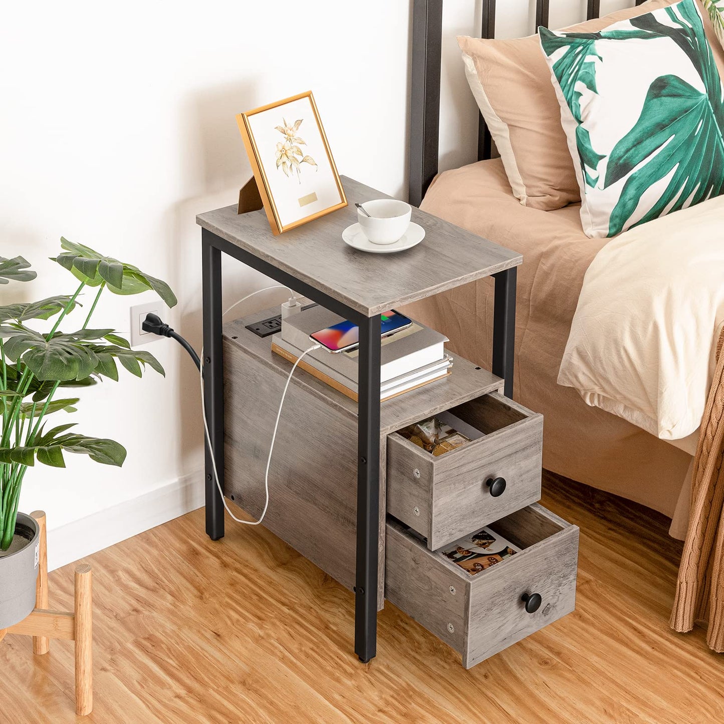 Gray side table with drawers next to a bed with decorative pillows and a plant.