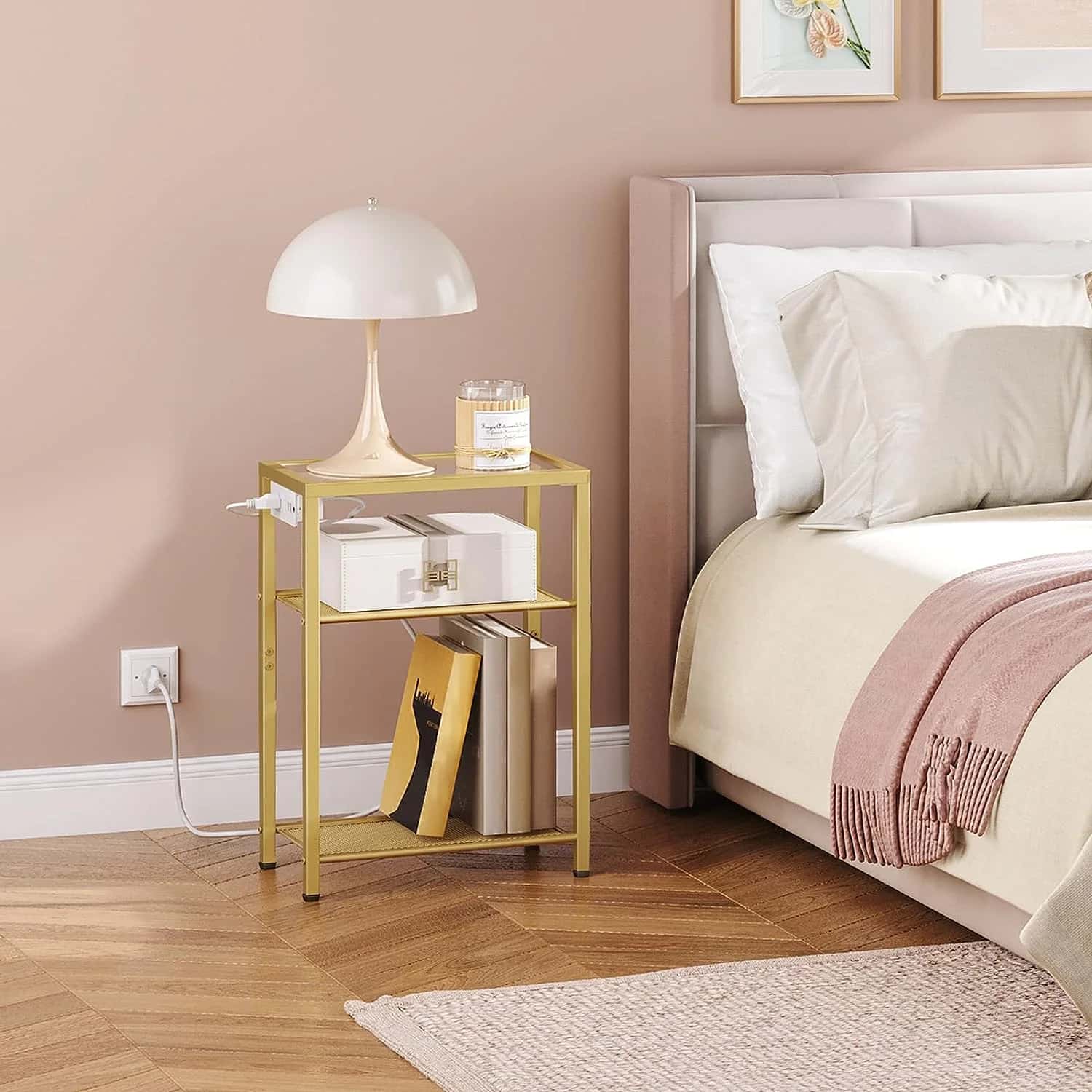 Gold nightstand with lamp and books next to a bed with pink blanket and pillows.