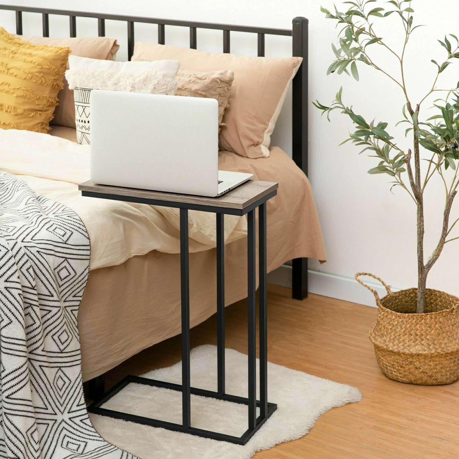 Small black metal side table with a laptop on it next to a bed with pillows and a plant in the corner.