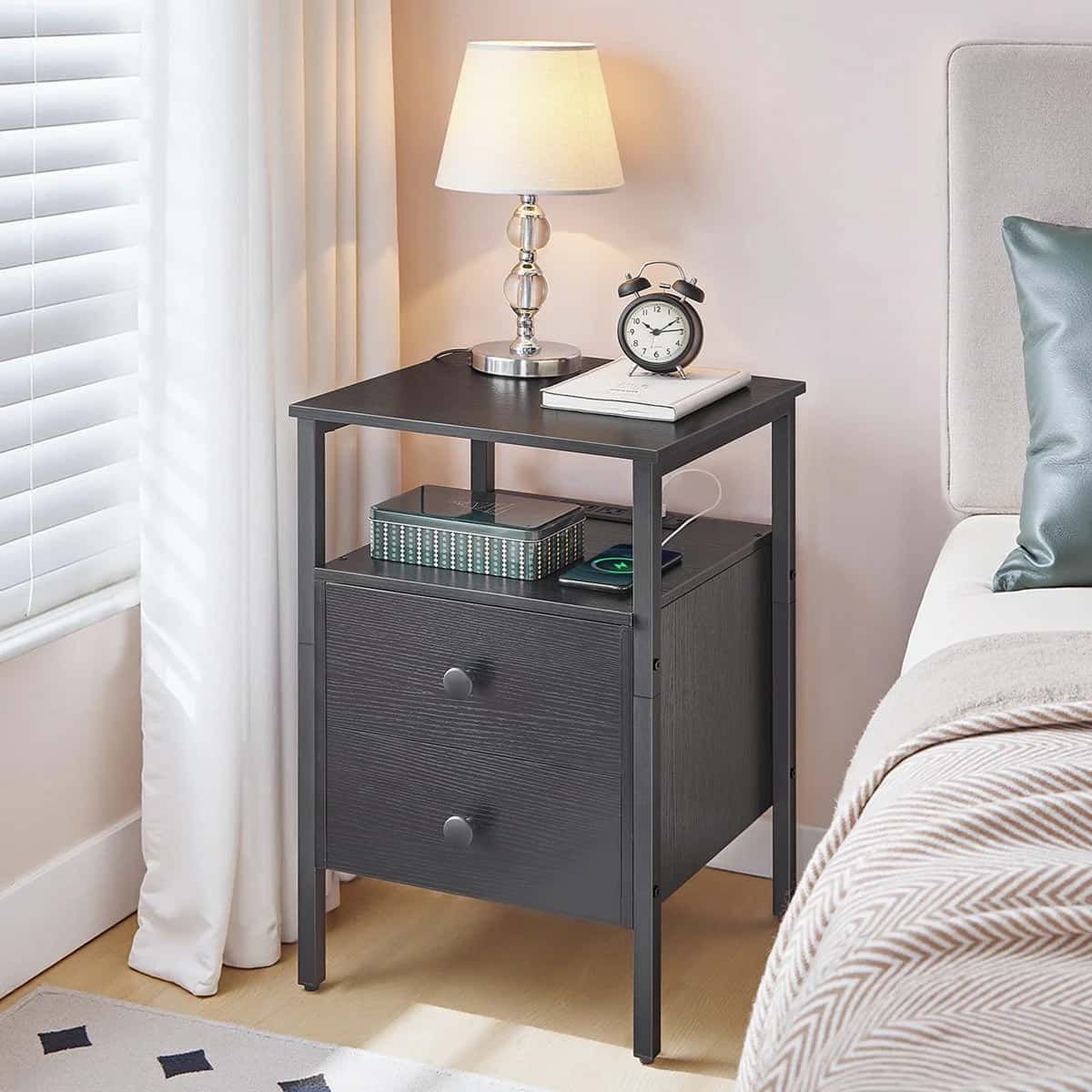 Black nightstand with lamp, clock, and books next to a bed in a bedroom.