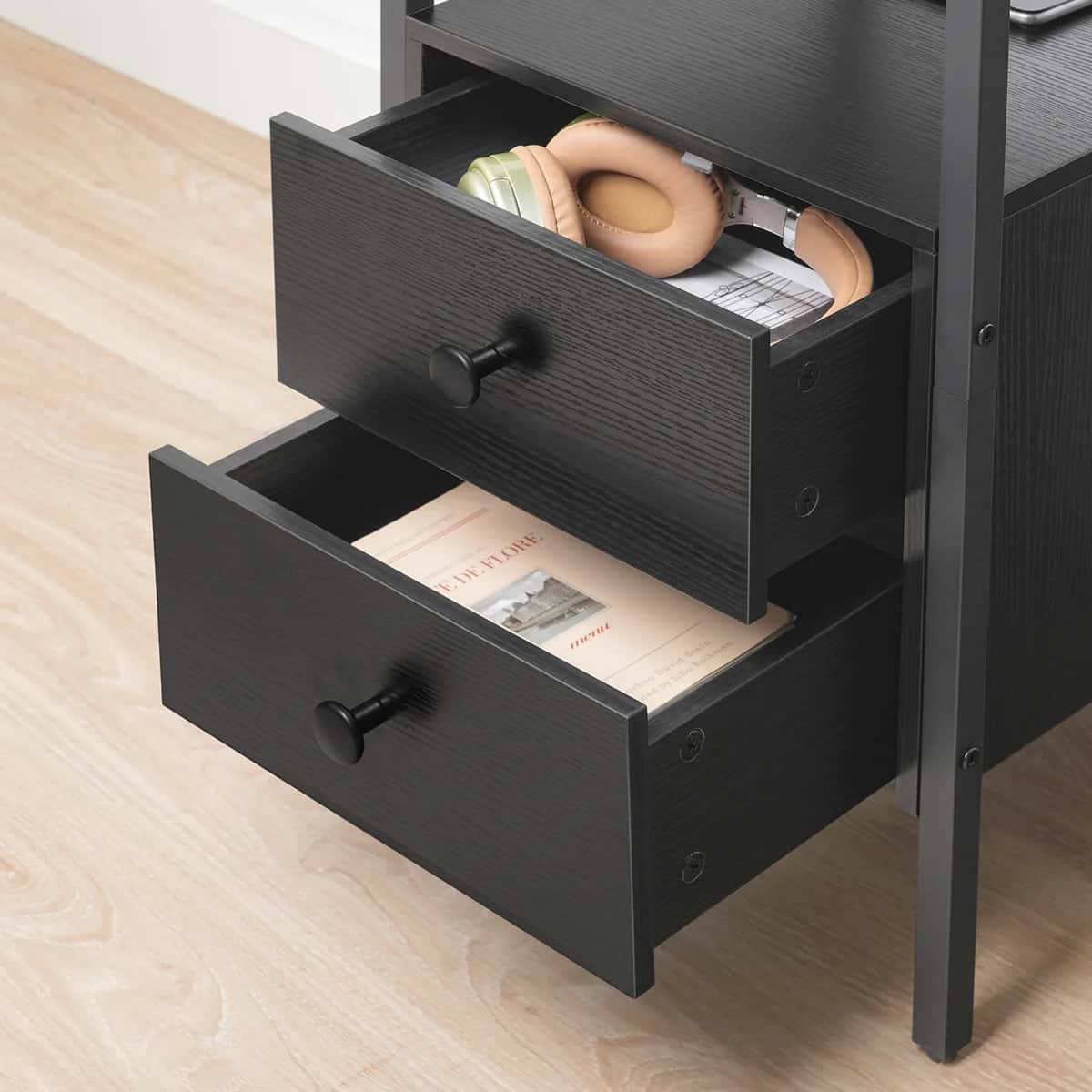 Black nightstand with open drawer containing books and a magazine on a wooden floor.