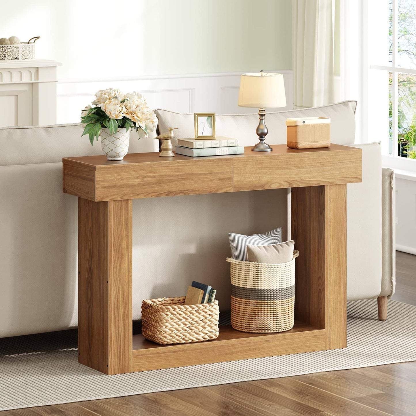 A light oak wood console table styled in a bright living room next to a cream sofa, decorated with a vase of white flowers, a table lamp, and storage baskets underneath.