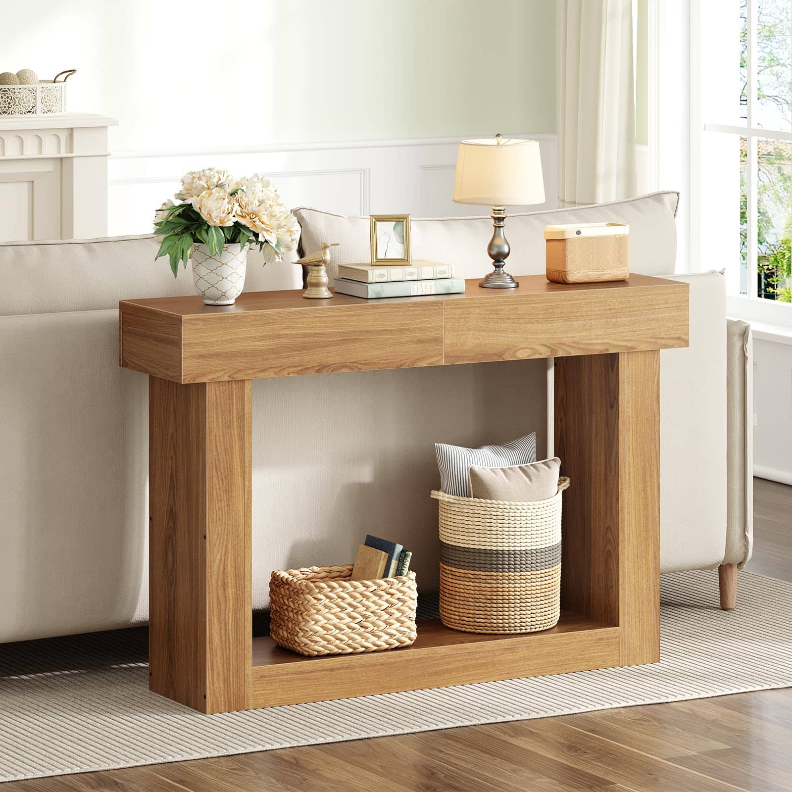A light oak wood console table styled in a bright living room next to a cream sofa, decorated with a vase of white flowers, a table lamp, and storage baskets underneath.