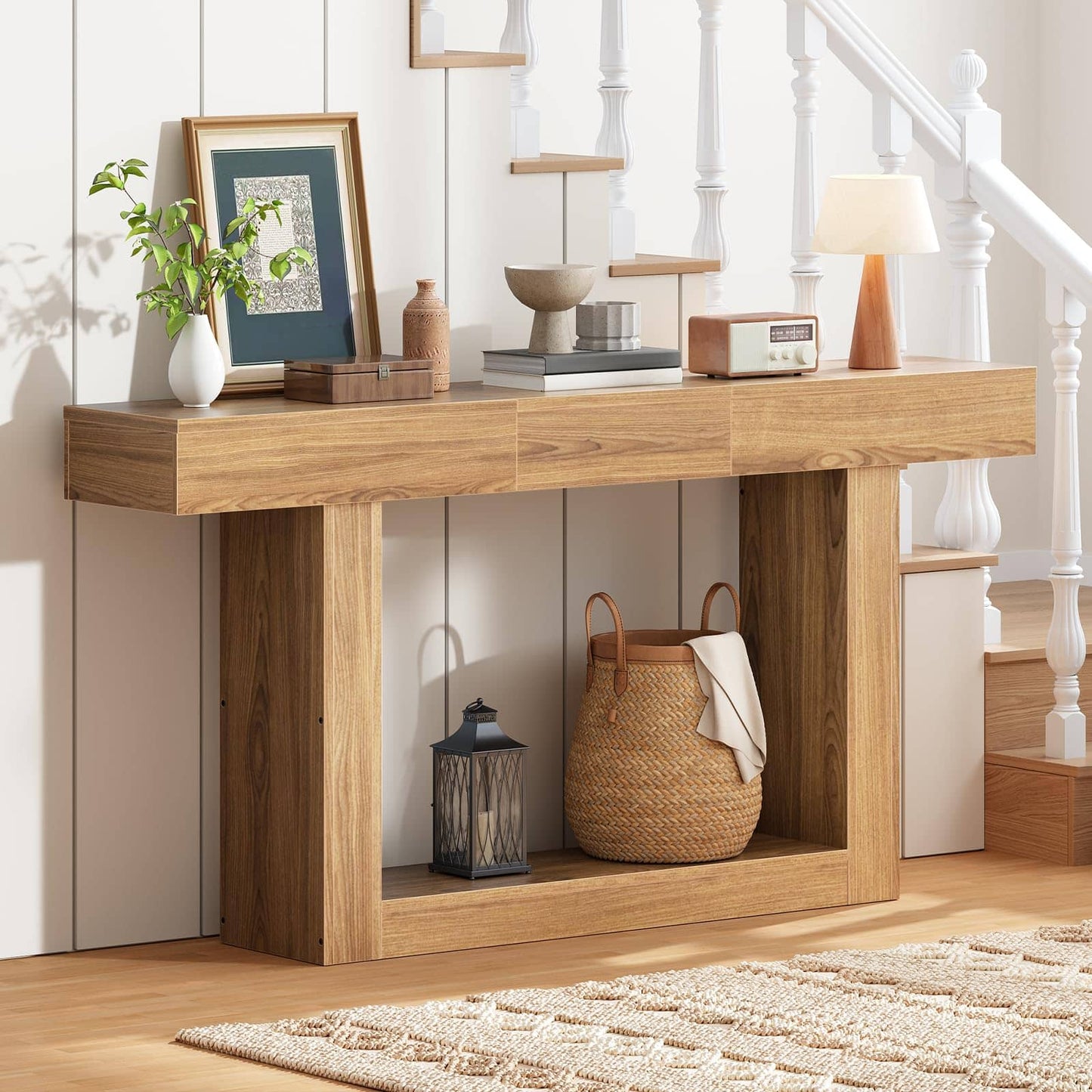 A light oak wood console table styled in a bright entryway with a white staircase, holding home decor items like a framed print, books, a small lamp, and a woven basket on its open lower shelf.