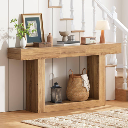 A light oak wood console table styled in a bright entryway with a white staircase, holding home decor items like a framed print, books, a small lamp, and a woven basket on its open lower shelf.