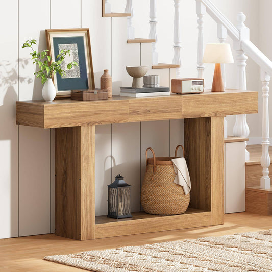 A light oak wood console table styled in a bright entryway with a white staircase, holding home decor items like a framed print, books, a small lamp, and a woven basket on its open lower shelf.