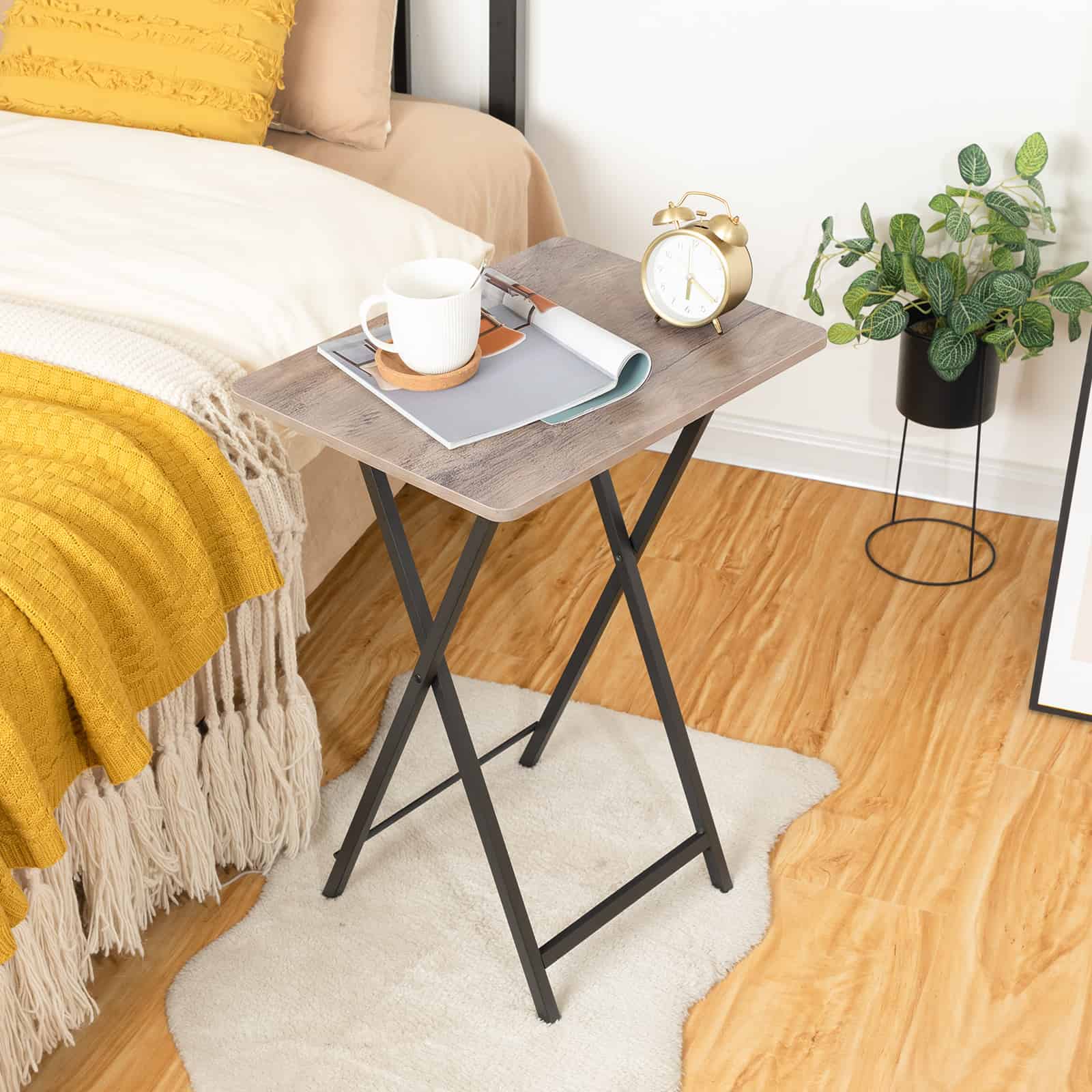 Industrial style grey wood folding side table used as a compact bedroom nightstand with a coffee cup and alarm clock.