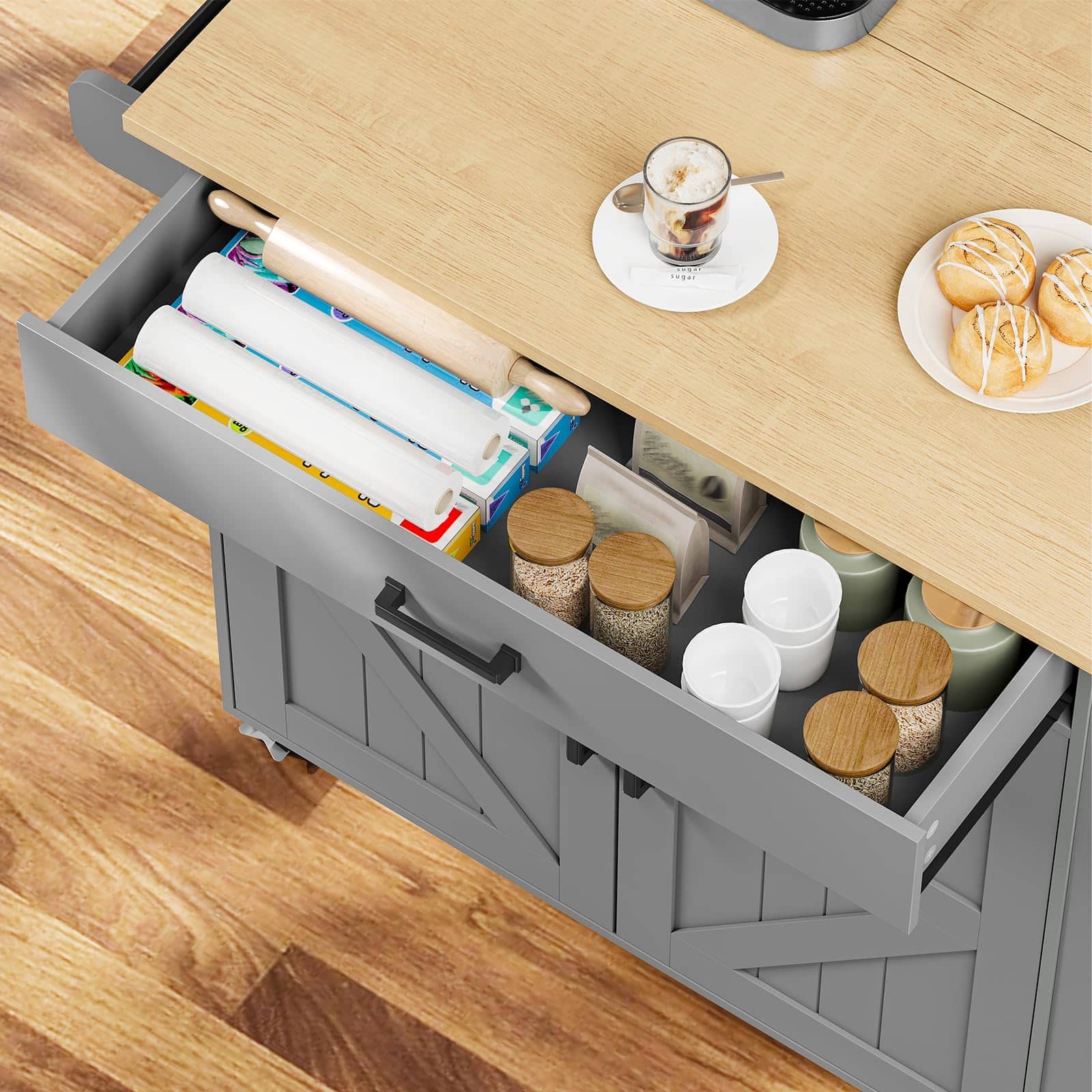 A gray rolling storage cabinet with a light wood top, shown with one drawer open to reveal storage bins, jars, and kitchen items. A plate of pastries and a cup sit on the tabletop, and the cabinet is placed on a wood floor.