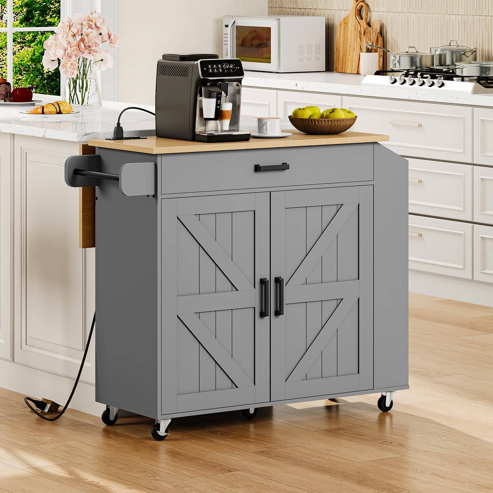 A gray kitchen cart with a light wood top and black caster wheels, shown in a bright, modern kitchen. The cart features two barn-style doors with decorative X-brace panels, a single drawer with a black handle, and a pull-out side arm.