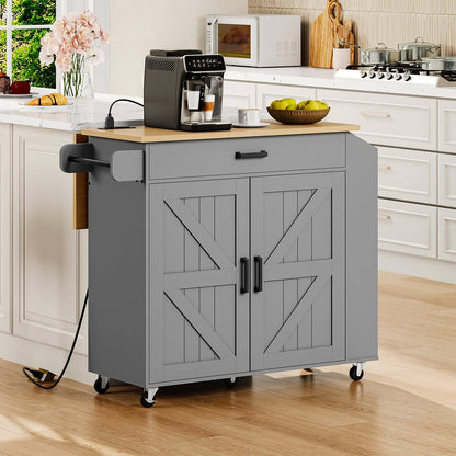 A gray kitchen cart with a light wood top and black caster wheels, shown in a bright, modern kitchen. The cart features two barn-style doors with decorative X-brace panels, a single drawer with a black handle, and a pull-out side arm.