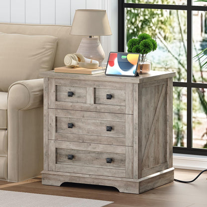 Rustic gray sofa end table with three drawers holding a white shaded lamp next to an armchair in a living room.