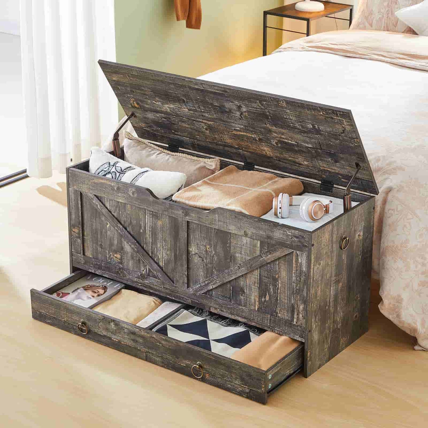 Rustic grey wood storage bench with open lid and bottom drawer holding blankets at the end of a bed.