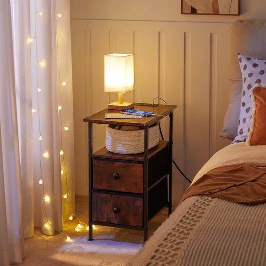 Nighstand with lamp and basket next to a bed in a bedroom with string lights on the curtain.