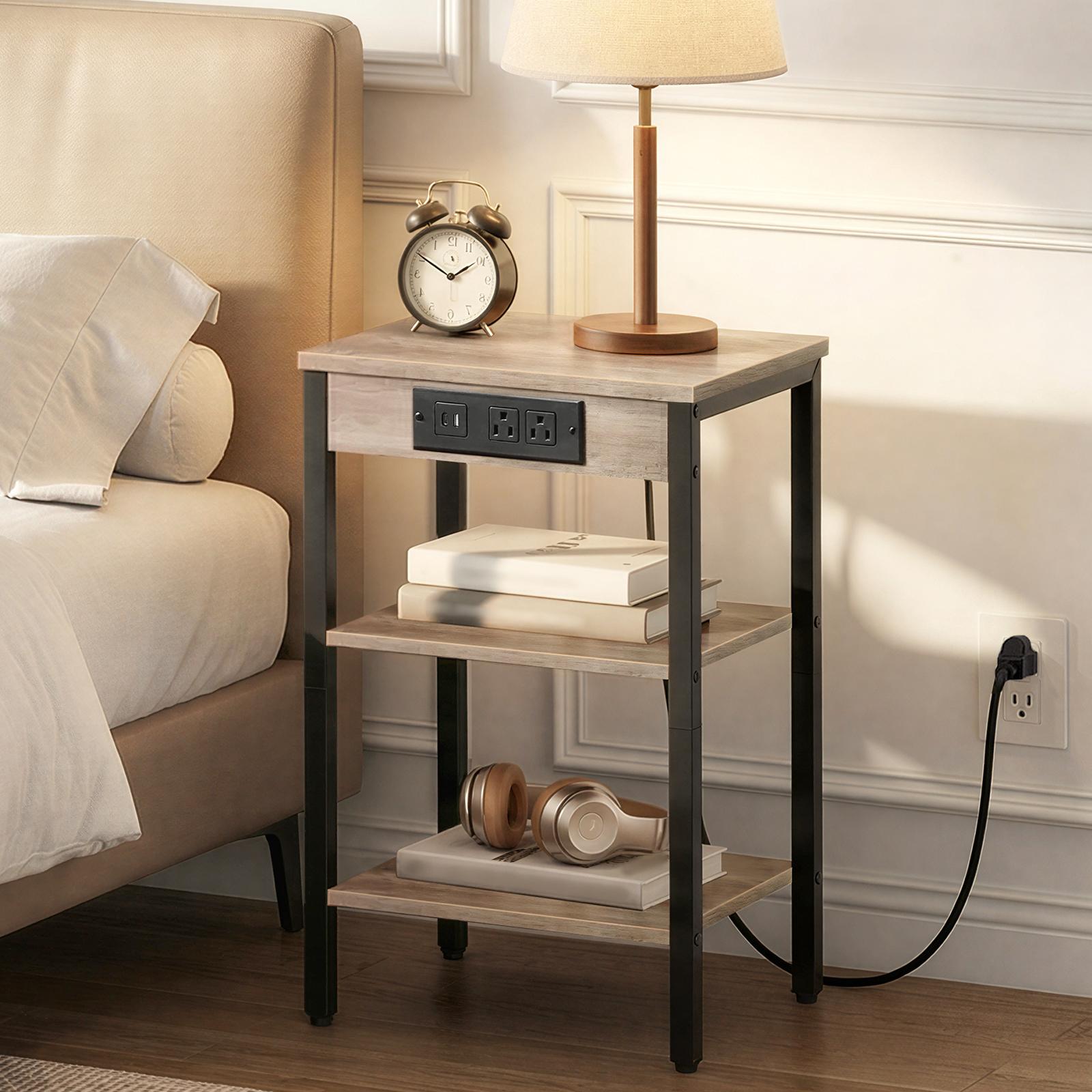 Slim wooden nightstand with power outlets placed next to a bed with an alarm clock and a lamp.